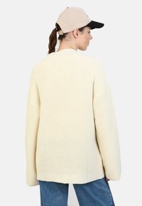 Kremowy, oversize'owy sweter z dzianiny o fakturowym wzorze, z długimi rękawami i luźnym krojem, połączony z jasnoniebieskimi dżinsami i beżową czapką.