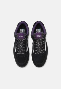Černé semišové tenisky s bílým bočním pruhem, texturovaným jazykem s fialovými a šedými vzory a tkanicemi. Logo Vans na jazyku a stélce.