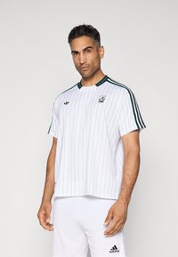 Mand iført en hvid Adidas sports t-shirt med tynde blå striber, sorte skulderstriber og matchende hvide Adidas shorts mod en ensfarvet baggrund.