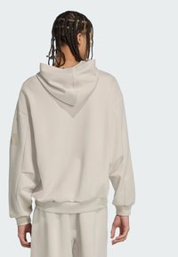 Hoodie overdimensionato beige con un ampio cappuccio, polsini e orlo a costine. Presenta un dettaglio del logo discreto sulla manica. Texture liscia.