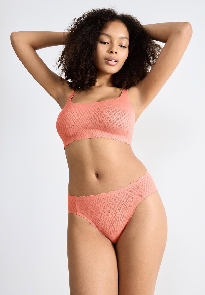 Donna con capelli ricci che indossa un reggiseno e un perizoma senza cuciture abbinati a un motivo corallo, in posa con le braccia sollevate dietro la testa su uno sfondo bianco.