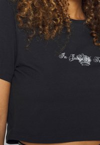 T-shirt noir court en coton avec des manches courtes. Arbore le texte blanc « In Juicy We Trust » et un graphique de couronne décoratif.