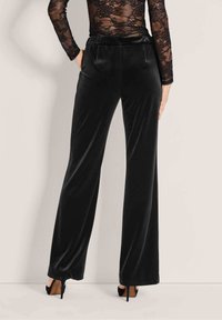 Vrouw in zwarte wijde fluwelen broek en een zwarte lange mouwen kanten top, staande tegen een effen lichte achtergrond.