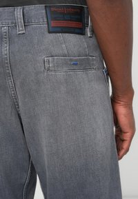 Jeans in denim grigio con una texture leggermente sbiadita, caratterizzati da un'etichetta nera con testo rosso e blu e un piccolo dettaglio di cucitura blu sulla tasca posteriore.