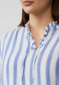 Gestreepte blouse in witte en blauwe stof, met een gerimpelde halslijn en een opening zonder knopen. De textuur lijkt licht en zacht te zijn.