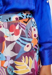 Satynowa niebieska bluzka zestawiona z kolorową, wzorzystą spódnicą z motywami kwiatowymi i abstrakcyjnymi w odcieniach fioletu, pomarańczu i zieleni morskiej.