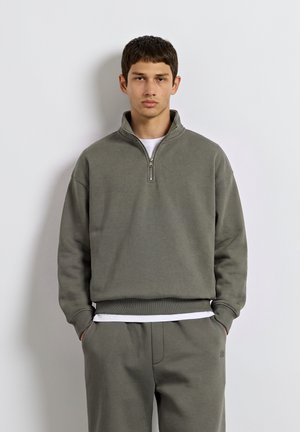 Ung mand med kort mørkt hår iført en olivengrøn sweatshirt med halv lynlås og matchende bukser, hænderne i lommerne.