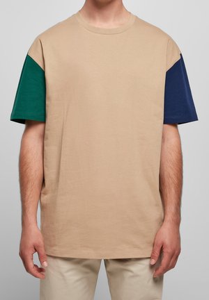 T-shirt beige à manches courtes avec une manche verte à gauche et une manche bleu marine à droite, porté par une personne debout.