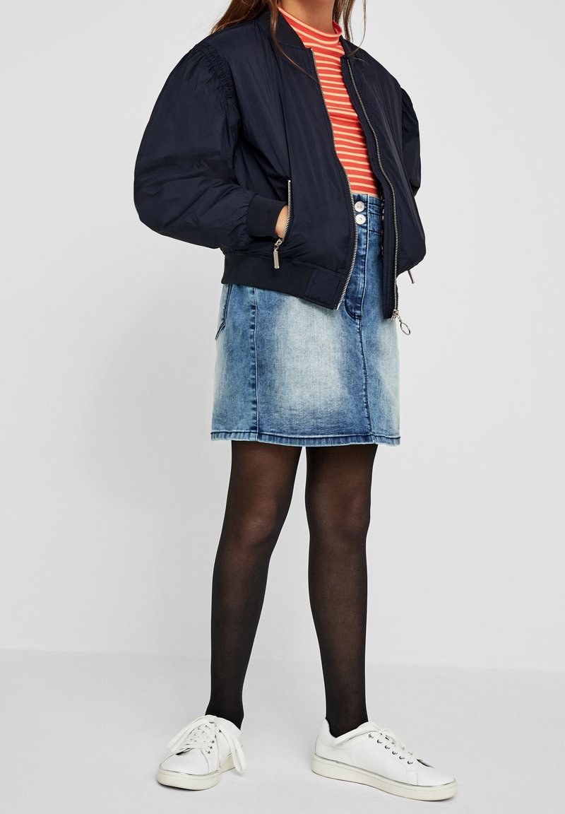 Personne portant une veste bomber bleu marine, un haut rayé rouge et blanc, une jupe en jean, des collants noirs et des baskets blanches, debout devant un fond uni.