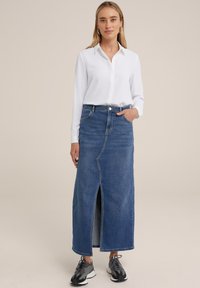 Falda maxi de mezclilla en azul oscuro con una abertura frontal, diseño de cinco bolsillos y presillas para el cinturón, combinada con una camisa blanca de botones y zapatillas.