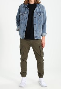 Light blue denim jacket over a black shirt, paired with olive cargo pants with side pockets and white sneakers. Plain background.