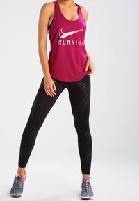 Magenta Nike tanktop met de tekst "RUNNING", gecombineerd met zwarte leggings met een grafisch patroon en grijze sportschoenen.