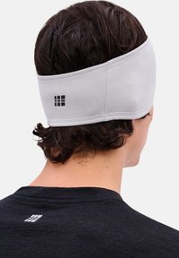 Grijze hoofdband gemaakt van gestructureerde stof, met een klein zwart logo. Draagt het achter kort, krullend haar met een zwarte shirt zichtbaar.