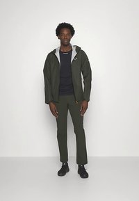 Veste imperméable vert foncé avec doublure gris clair, fermeture éclair à l'avant et capuche ajustable. Associée à une chemise bleu marine et un pantalon slim assorti.