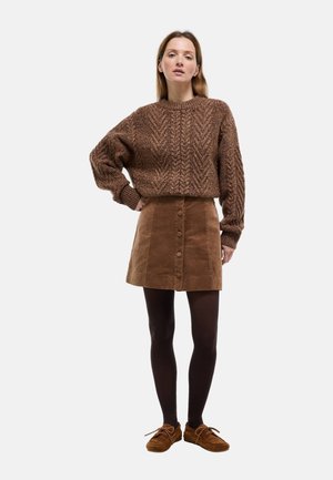 Pull en maille texturée marron avec un col rond, associé à une jupe en velours côtelé marron à boutons sur le devant et des collants noirs, complété par des chaussures marron.