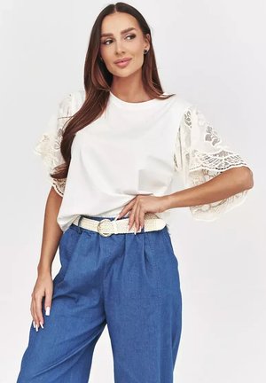 Kobieta w białej bluzce z koronkowymi rękawami, niebieskich spodniach z wysokim stanem i białym plecionym pasku, pozująca z jedną ręką na biodrze.