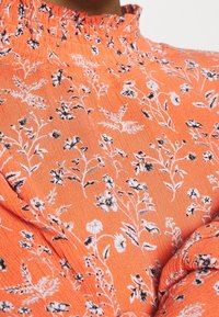 Blouse orange à col montant avec imprimé floral blanc et noir et détails de tissu froncé au niveau du col et des manches.