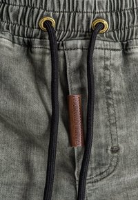 Pantalones de jogging verdes con cintura ajustable por cordón, con cordón negro y ojetes dorados, y una etiqueta de cuero marrón con texto en relieve.