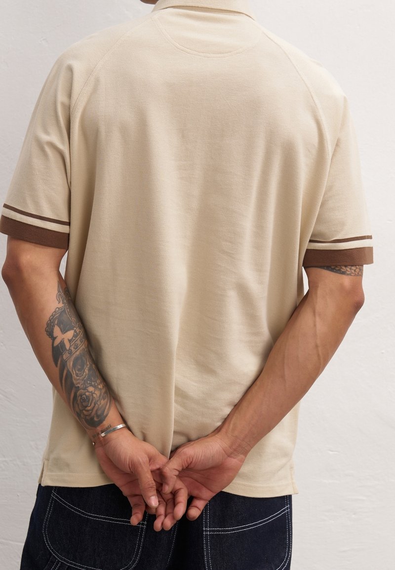 Polo beige avec des manches raglan courtes. Accents rayés marron sur les manches. Tissu doux, coupe décontractée et col classique.