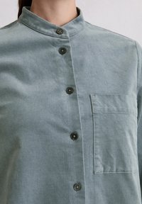 Lichtgrijs knoopshirt met een staande kraag en een borstzak, gedragen door een persoon met het haar in een staart.