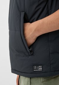 Schwarze Pufferjacke mit einer Reißverschlusstasche, einem Etikett mit Pflegehinweisen und einer glatten, matten Textur.