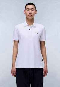 Giovane uomo che indossa una maglietta polo bianca con un piccolo logo e pantaloni neri, in piedi contro uno sfondo grigio chiaro uniforme.
