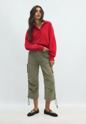 Femme debout portant une chemise rouge à manches longues, un pantalon cargo large et court vert olive, et des chaussures plates noires sur un fond uni.
