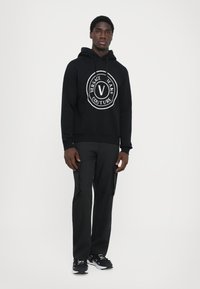 Mustmustrit, millel on ümmargune valge logo ja tekst "VERSACE JEANS COUTURE". Kombineeritud mustade cargo-pükstega ja mustade tossudega.