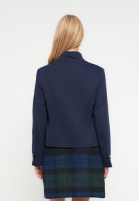 Στρατιωτικό μπλε, ραφτό σακάκ ι με γιακά με εγκοπές, εφαρμοστή γραμμή και ασημένια κουμπιά στα μανίκια, συνδυασμένο με καρό φούστα σε πράσινο και μπλε χρώμα.