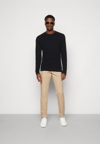 Black knitted sweater, khaki pants, and white sneakers. Accessories include square sunglasses. Model stands against a plain background.
