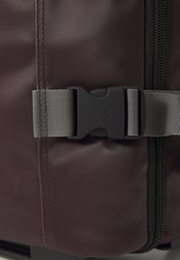 Dark purple travel bag with a smooth, matte finish, featuring a grey buckle strap and black zipper. Textured stitching along the seam.