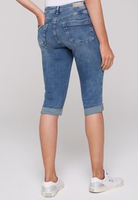 Jeans capri de denim azul claro con puños doblados, bolsillos traseros y una textura suave. Llevados con zapatillas blancas.