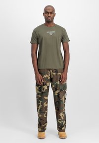Tricou scurt de culoare verde măsliniu, cu text imprimat, asortat cu pantaloni cargo camuflați, dotati cu multiple buzunare, completat cu cizme de culoare bej.