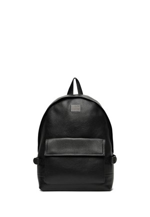 Czarny skórzany plecak z przednią kieszenią na klapę, górnym uchwytem i regulowanymi paskami bocznymi na białym tle.