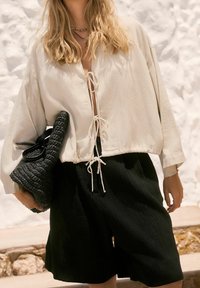 Lichtbeige oversized blouse met een strik bij de halslijn, gecombineerd met zwarte textuurshorts. Het model houdt een geweven zwarte handtas vast.