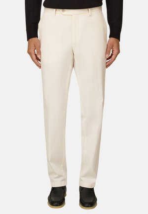 Homme portant un pantalon ajusté de couleur crème, un haut noir à manches longues et des chaussures noires avec des semelles marron clair, debout devant un fond neutre.