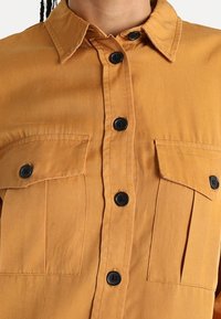 Knappt skjorta i ljusbrunt tyg med två bröstfickor, svarta knappar och klassisk krage. Mjuk textur, rak skärning.