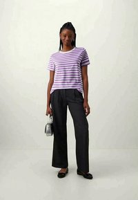 Camiseta a rayas moradas y blancas, de manga corta, combinada con pantalones negros de cintura alta. La modelo sostiene un pequeño bolso de mano plateado, lleva zapatos planos negros.