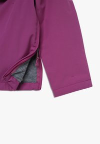 Lila jacka med ett mjukt yttermaterial. Har en zippered ficka och grå fleecefoder synligt vid öppningen.
