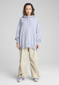 Hoodie surdimensionné bleu clair avec poche avant, manches longues et cordons de serrage, associé à un pantalon beige ample et des baskets avec des accents violets.