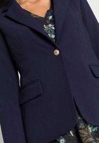 Blazer blu navy con un design strutturato, chiusura a un singolo bottone dorato e due tasche frontali. Il tessuto appare liscio e elegante.