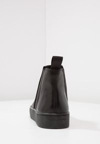 Botín de piel negra con paneles laterales elásticos, una pestaña para tirar y una suela texturizada. Presenta un diseño elegante y un branding minimalista.