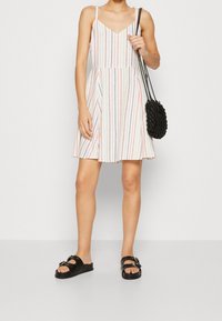 Gestreiftes ärmelloses Kleid in Weiß, Rot und Blau, mit enganliegendem Oberteil und ausladendem Rock. Kombiniert mit schwarzen Sandalen und einer strukturierten schwarzen Tasche.