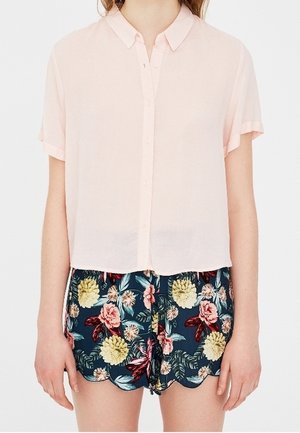 Kobieta ubrana w jasnoróżową koszulę z krótkim rękawem na guziki oraz granatowe kwieciste spodenki z falowanym obszyciem.