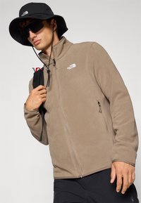 Brown fleece jacket with a high collar, front zipper, and logo on the chest. Worn with a black wide-brim hat and sunglasses.