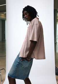 Kortærmet, lyserød ternet skjorte med en afslappet pasform, parret med blå denimshorts. Skjorten har en knaplukket krave og lommer.