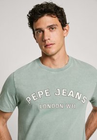 Žalias medvilnės marškinėliai su užrašu "PEPE JEANS LONDON W11" kreivuoju, ryškiu šriftu. Klasikinis apvalus iškirpimas ir trumpomis rankovėmis.