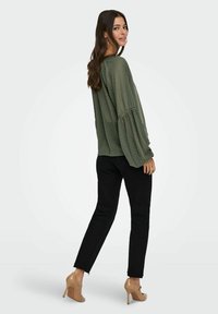 Donna con lunghi capelli castani, che indossa una blusa verde oliva a trama, jeans neri e tacchi beige, in una posa di tre quarti di spalle.