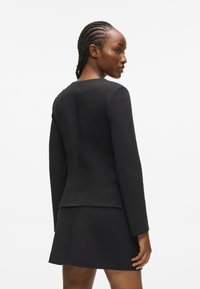 Zsinóros hajú nő egy testhez simuló fekete hosszú ujjú felsőt és ehhez illő rövid szoknyát visel, egy sima fehér háttér előtt áll.