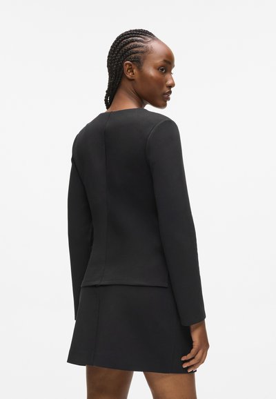 Femme aux cheveux tressés portant un haut ajusté à manches longues noir et une jupe courte assortie, se tenant devant un fond blanc uni.